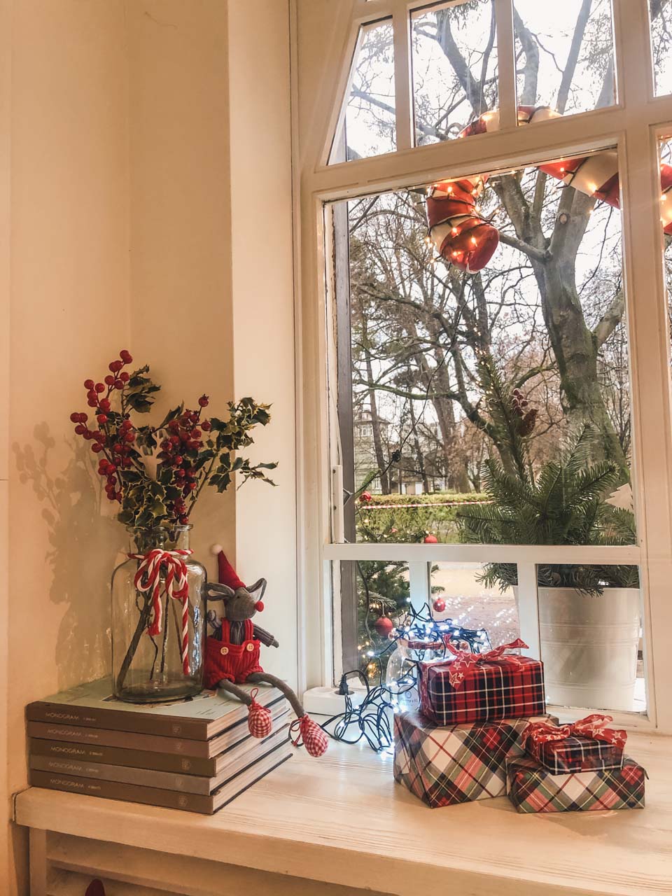A couple of wrapped presents, a stack of books and a vase with holly on a window sill