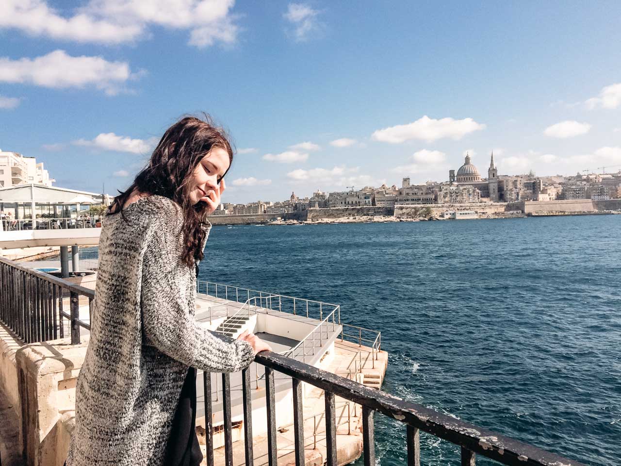 A girl standing on the promenade in Sliema, Malta with the Valletta skyline in the background
