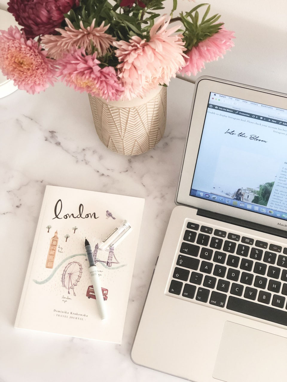 A travel journal, a vase with fresh flowers and a laptop on a marble desk
