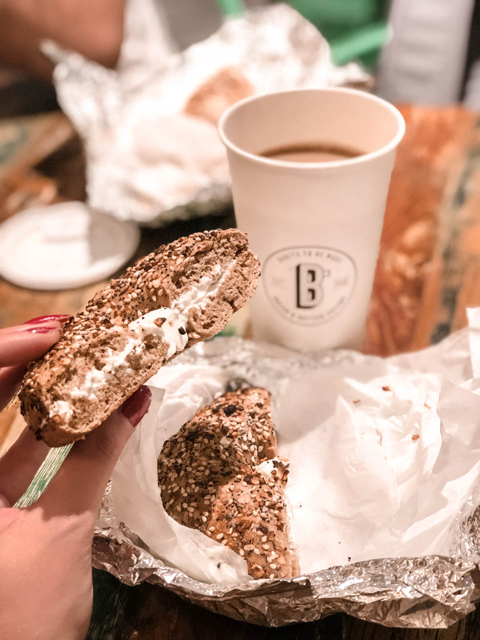 A girl holding a bagel with cream cheese with a cup of coffee in the background