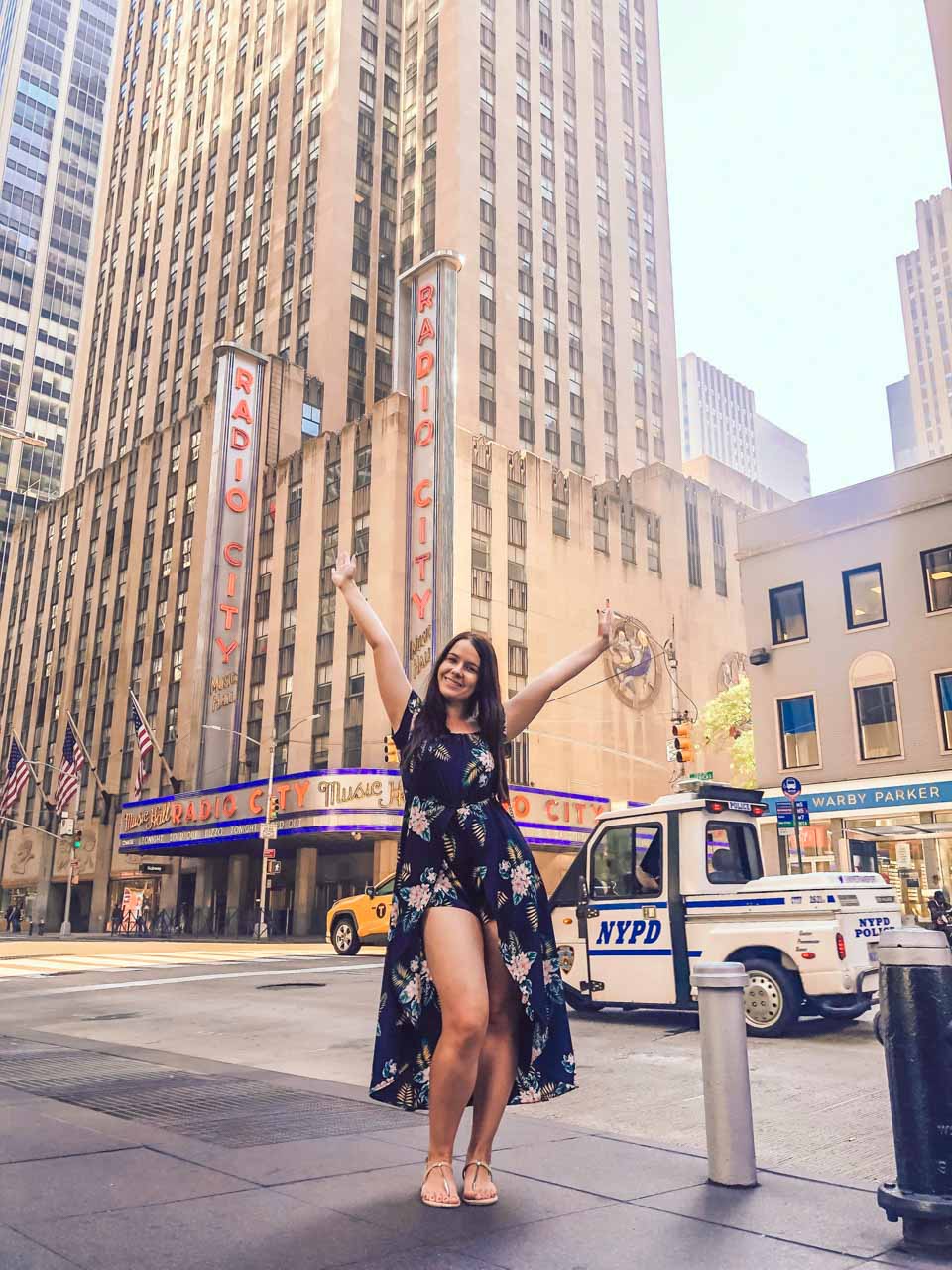 A happy girl in a floral dress throwing her hands up outside the Radio City Music Hall building