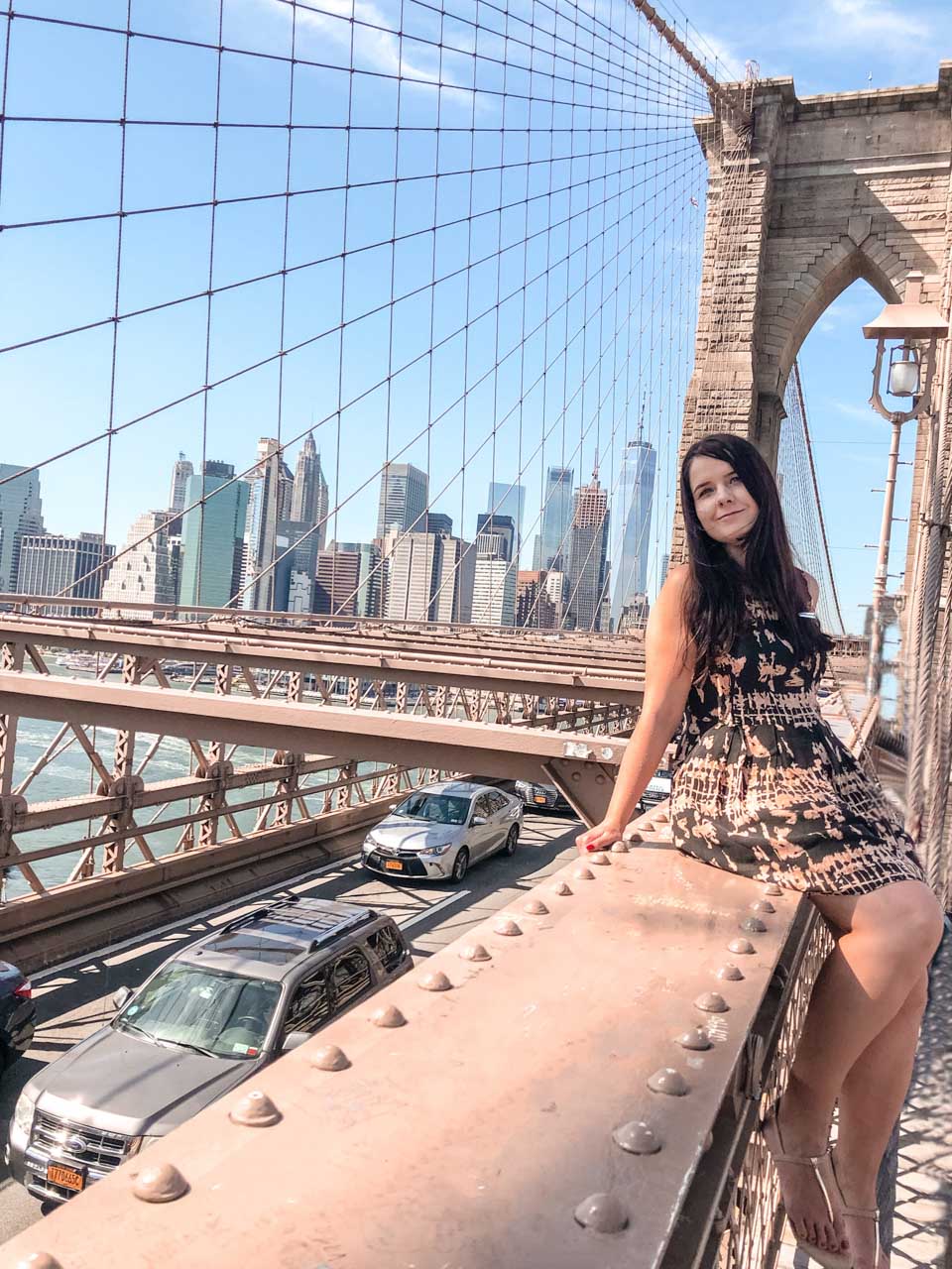 A girl in a dress sitting on the Brooklyn Bridge with the Manhattan skyline in the background