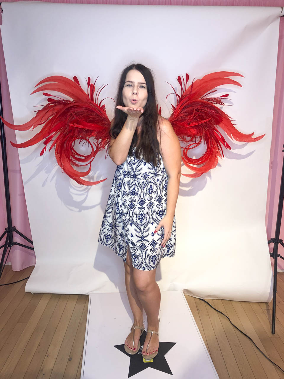 A girl in a patterned dress posing with the Victoria's Secret angel wings and blowing a kiss towards the camera