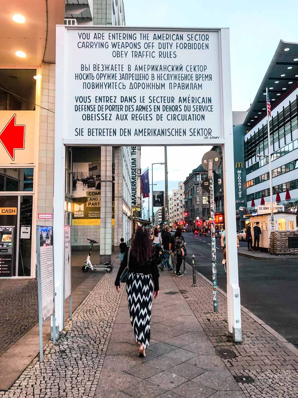 A girl in a maxi dress walking under the famous sign at Checkpoint Charlie in Berlin that says "You are entering the American sector" in various languages