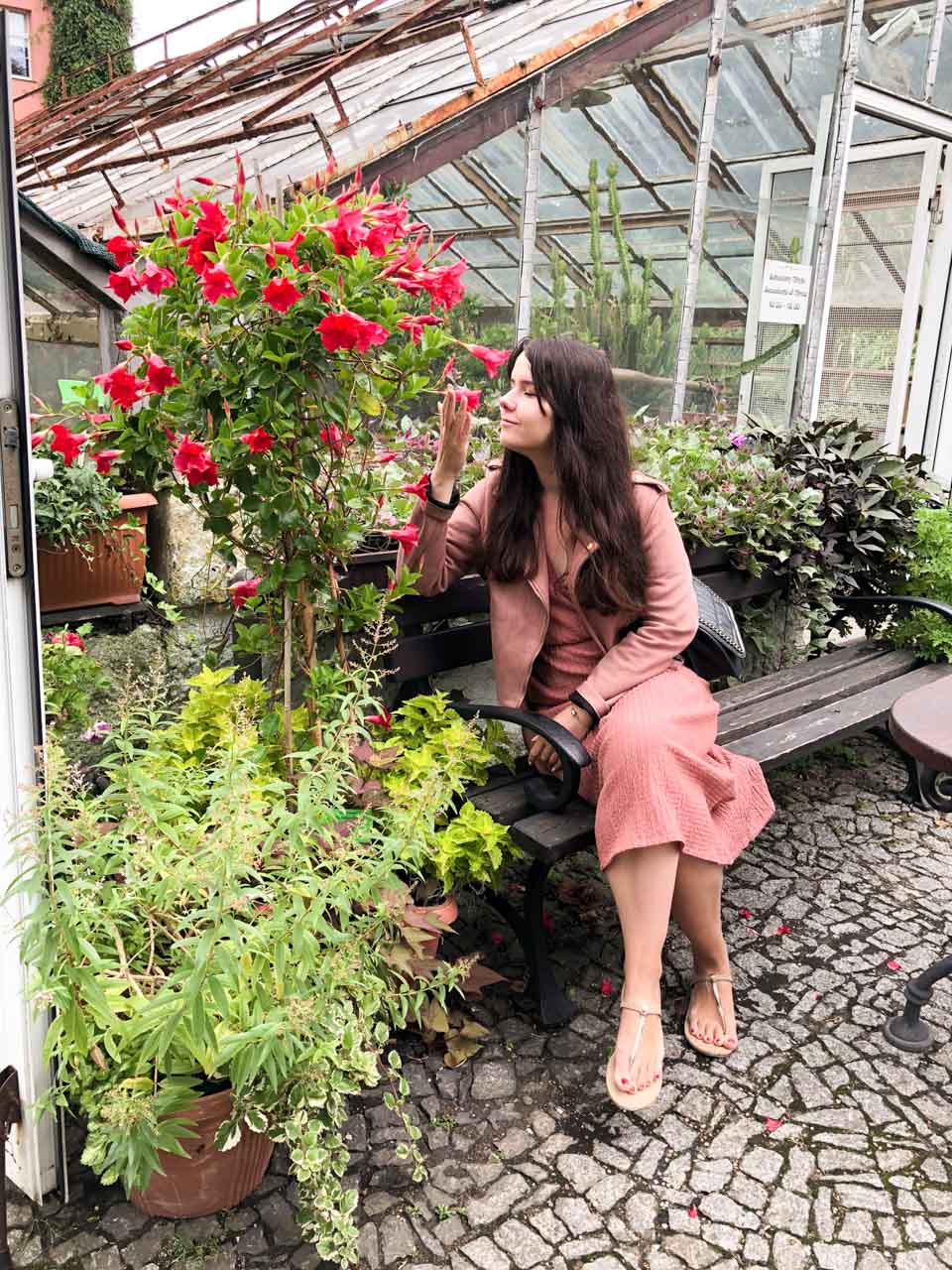 A woman in a pink dress sitting on a bench outside a greenhouse, smelling a red flower