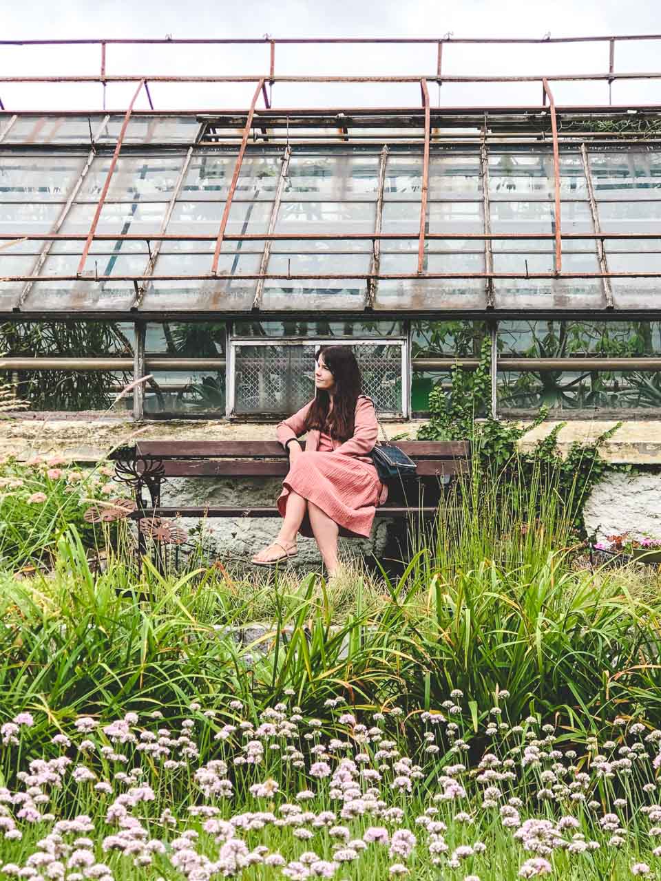 A woman in a pink dress looking to the side as she sits on a bench in front of a greenhouse