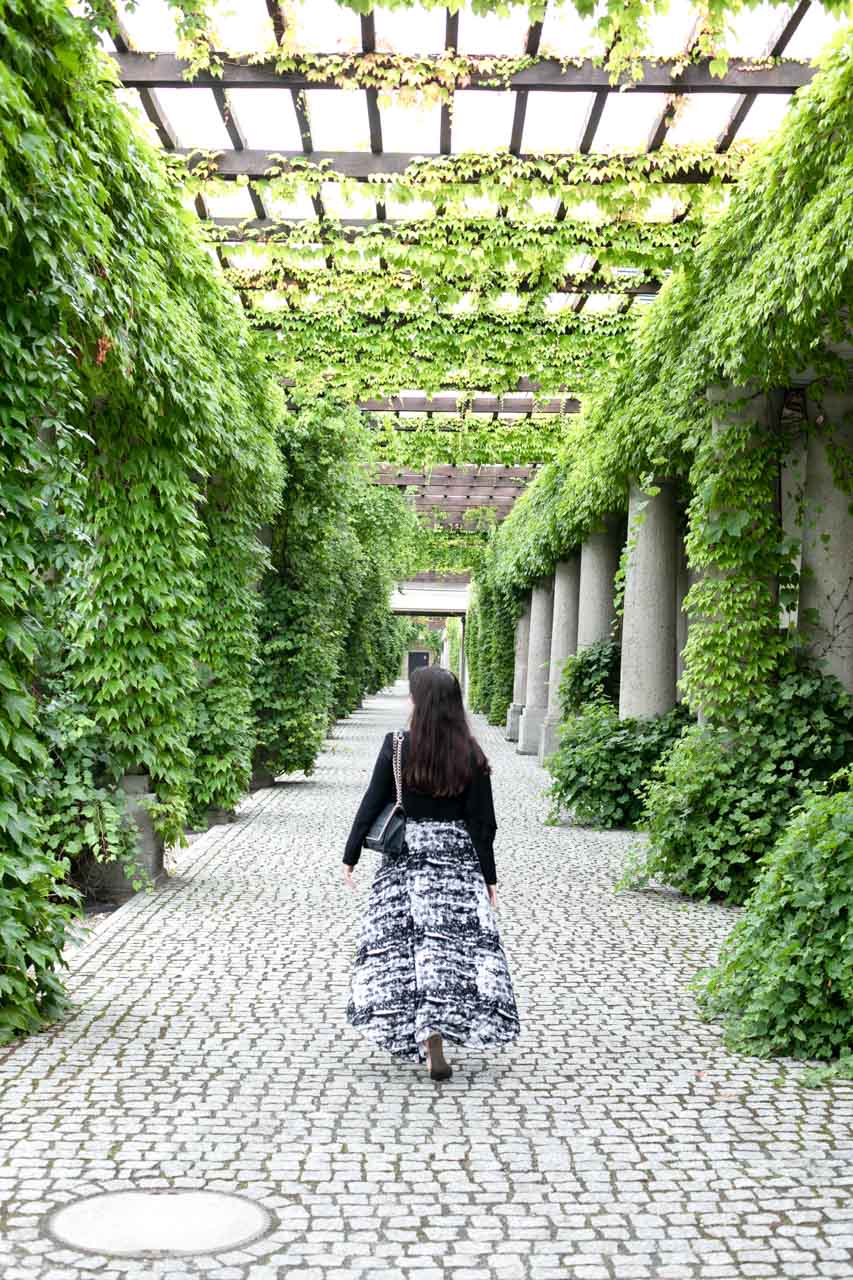 A woman in a black and white maxi dress walking under an ivy-covered pergola