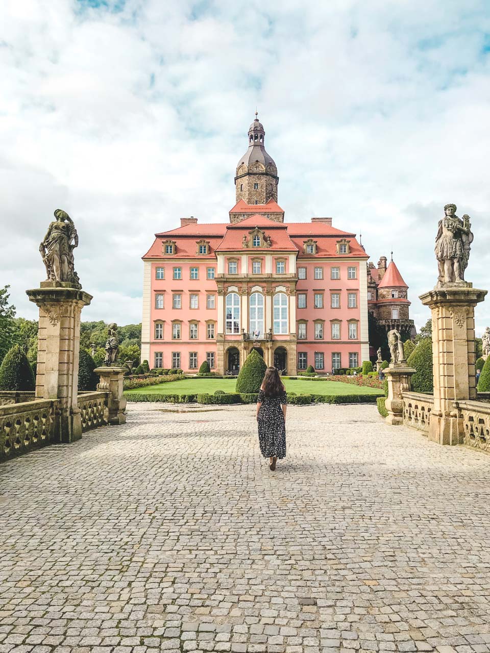 A woman in a black off-the-shoulder dress standing on the stone bridge leading to Książ Castle