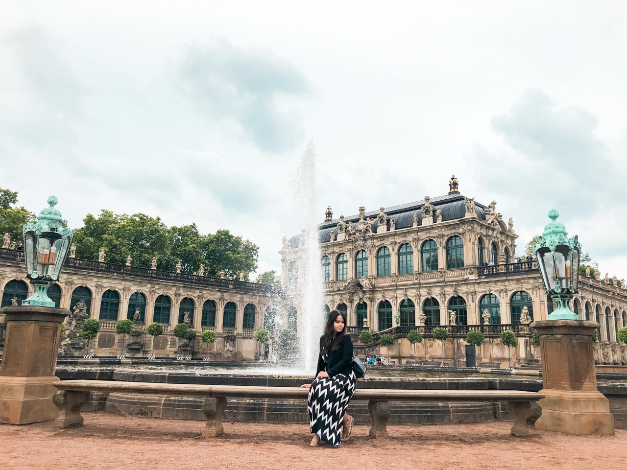 A woman in a maxi dress sitting on a bench in front of a fountain on Zwinger Square in Dresden