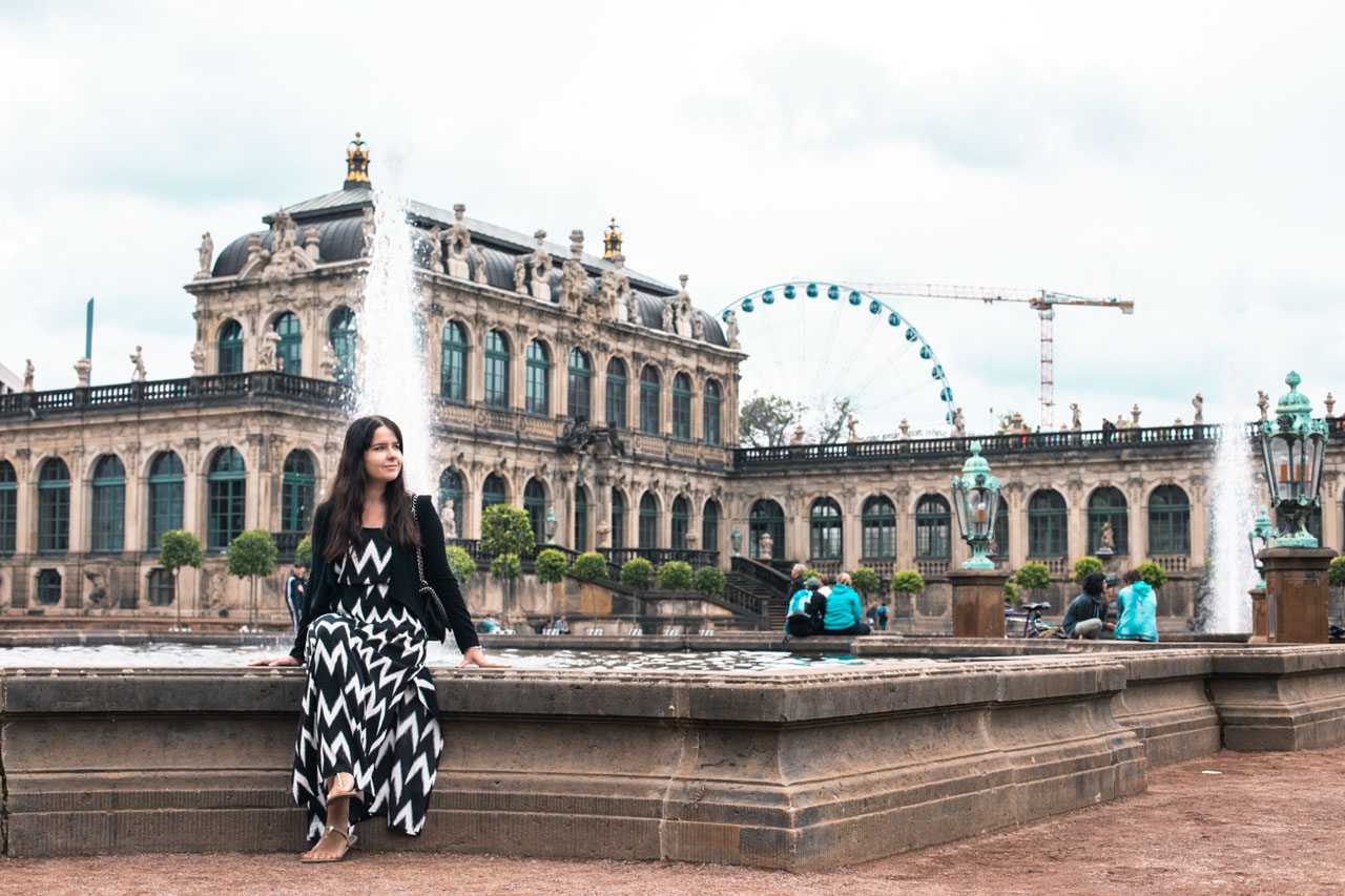 A dark-haired woman in a black and white maxi dress sitting on a fountain ledge on Zwinger Square