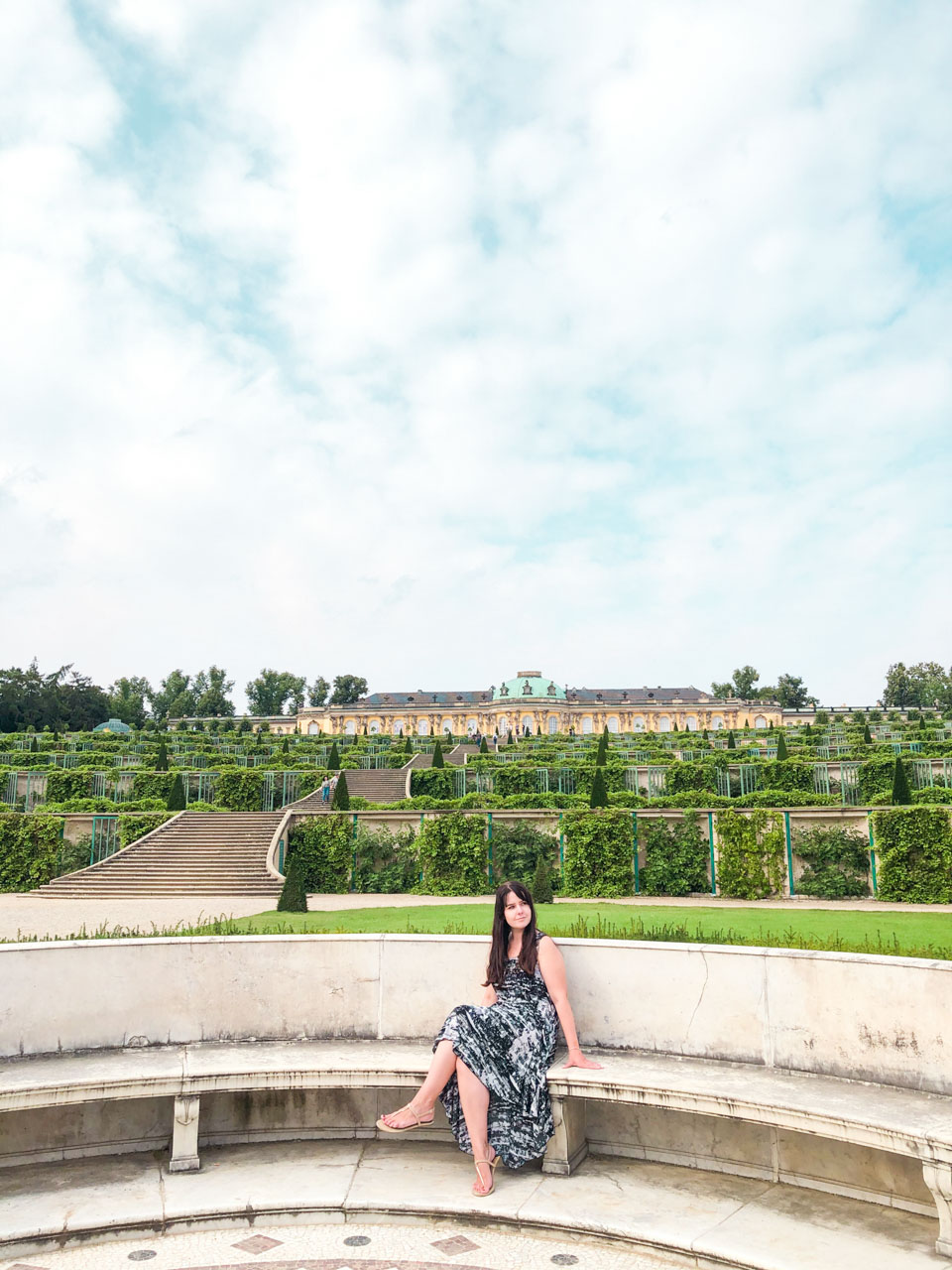 A woman in a black and white maxi dress sitting on a bench with the Sanssouci Palace behind her