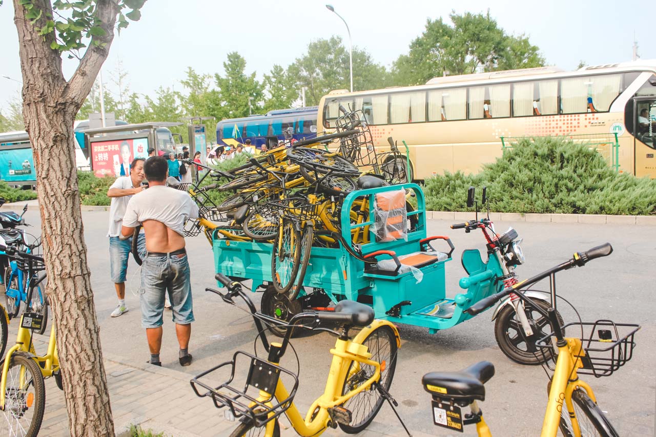 Two Chinese men standing next to a tricycle with a high pile of bicycles on it