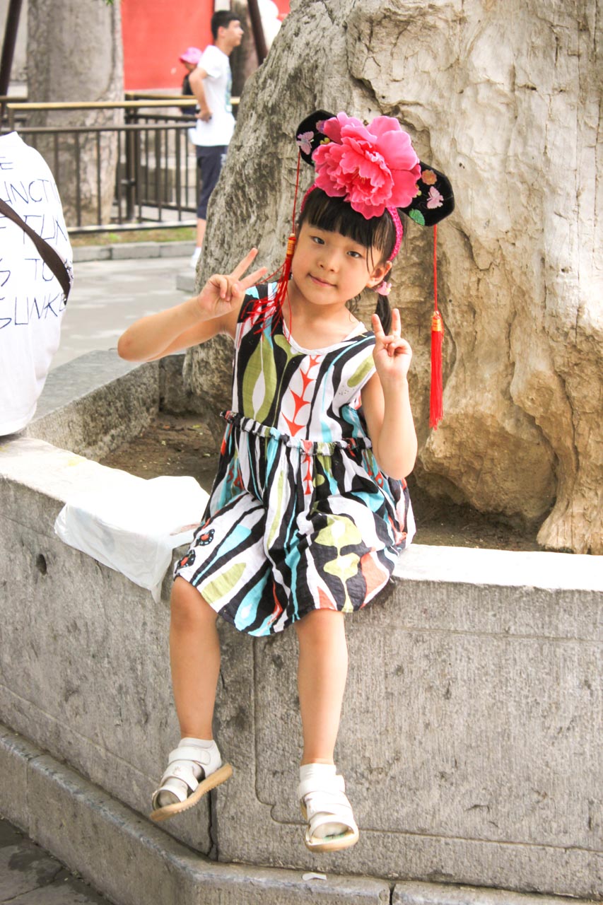 Young Chinese girl in a Chinese flower headpiece sitting on a stone ledge and throwing peace signs