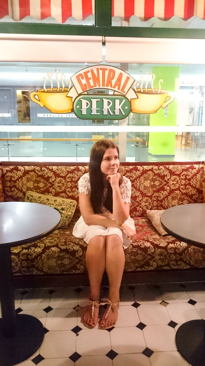 Girl in a white dress holding up her chin as she is sitting on a sofa inside the Central Perk café