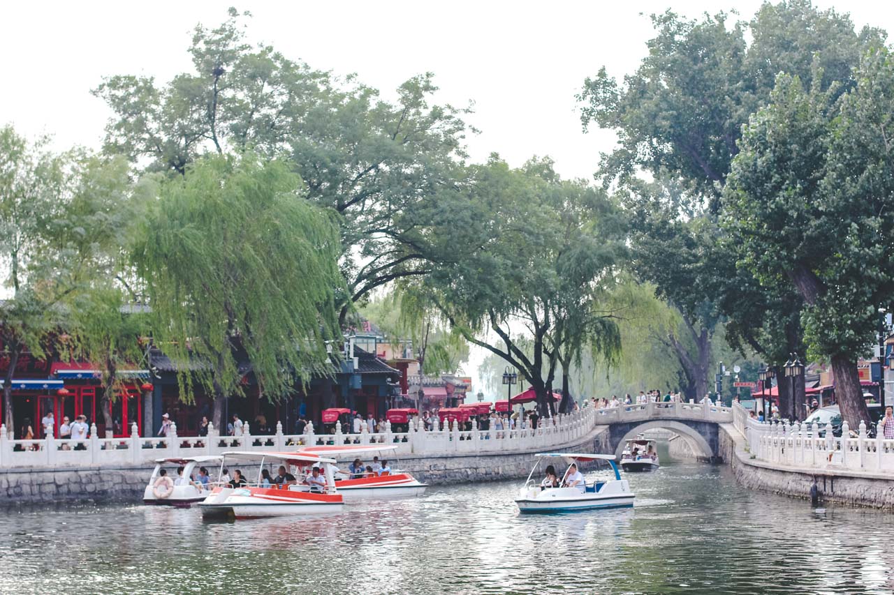 Pedalo boats passing under a stone bridge separating two alleys lined with shops and restaurants