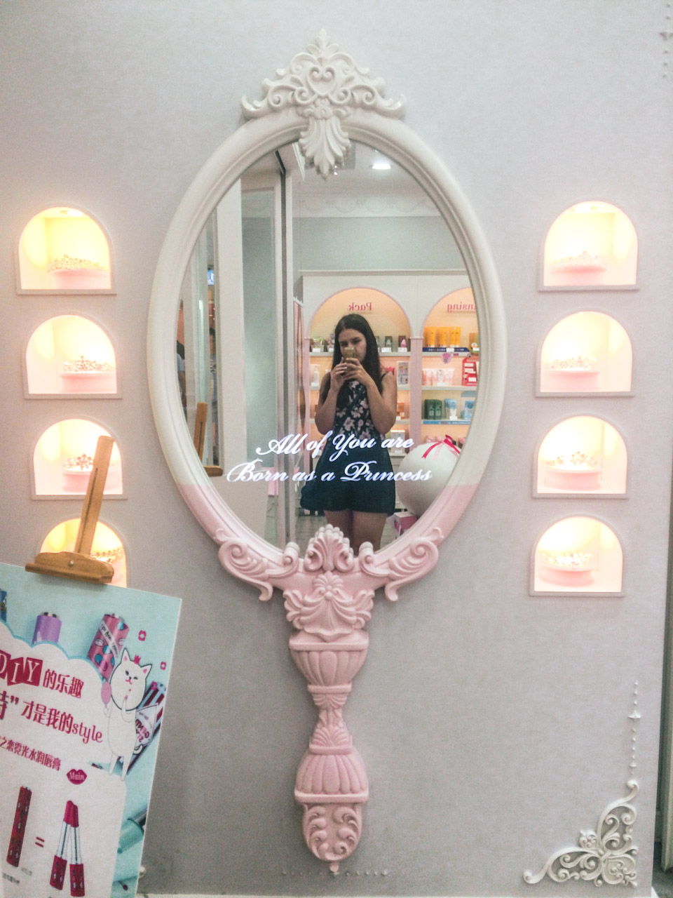 A dark-haired woman taking a selfie in a giant pink hand mirror hanging on a wall