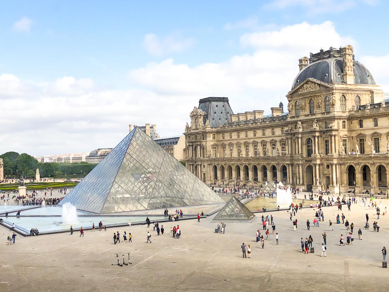 People walking around the Louvre Pyramid with the arcades housing Le Café Marly on the right side