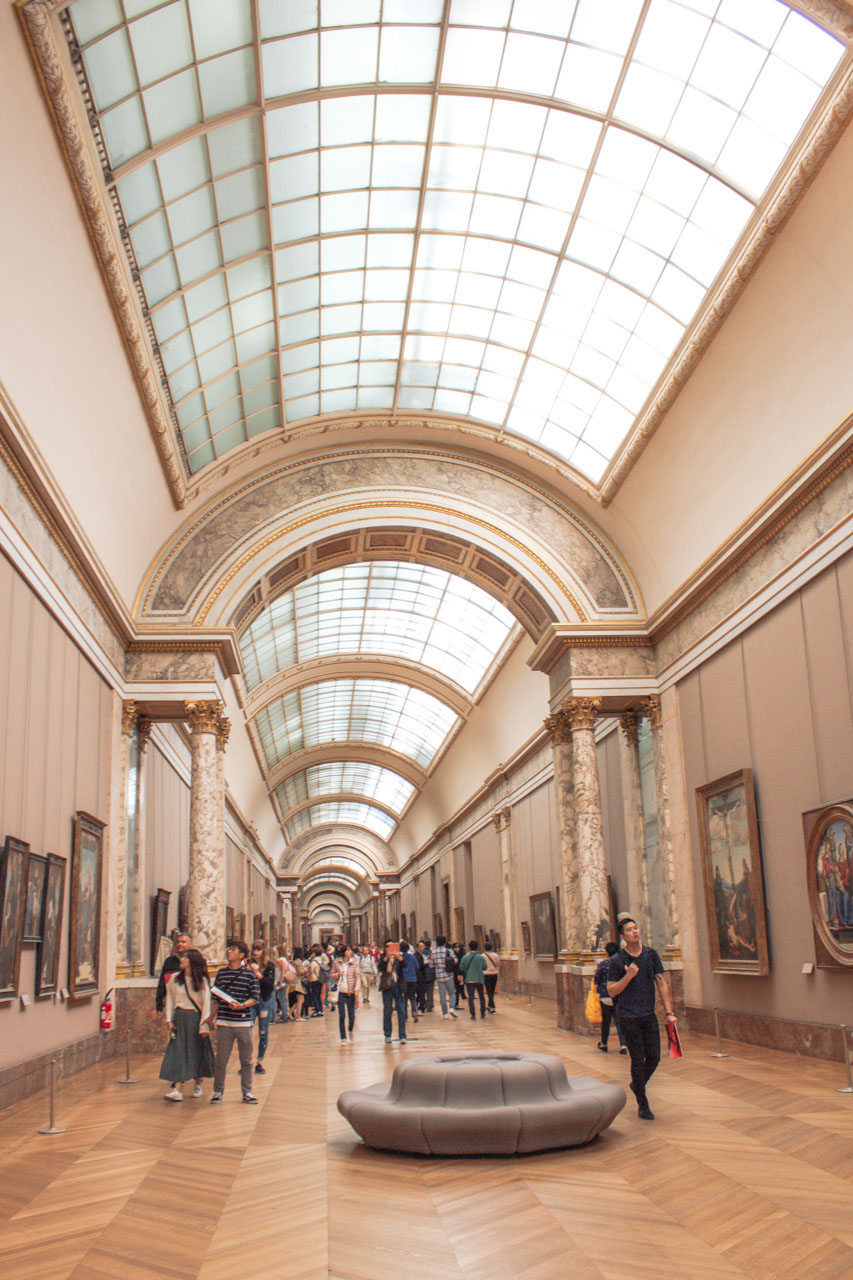 People wandering through the hallways and looking at the paintings on the walls in the Louvre Museum