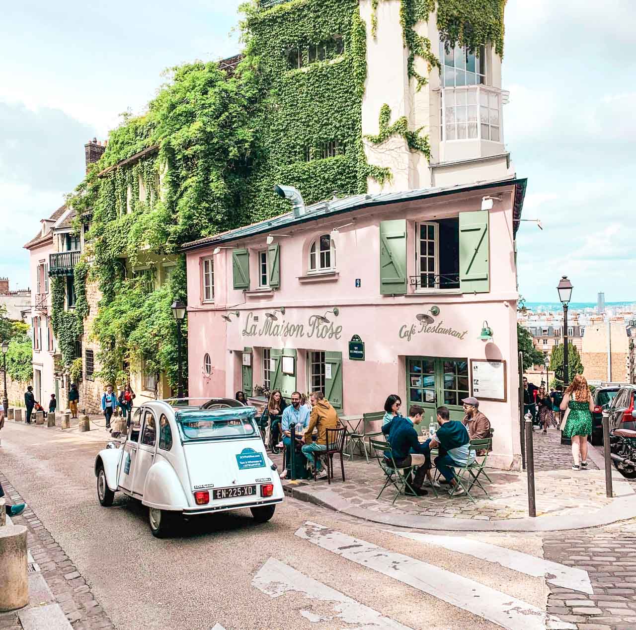 A white vintage car driving past people sitting outside La Maison Rose in Montmartre, Paris