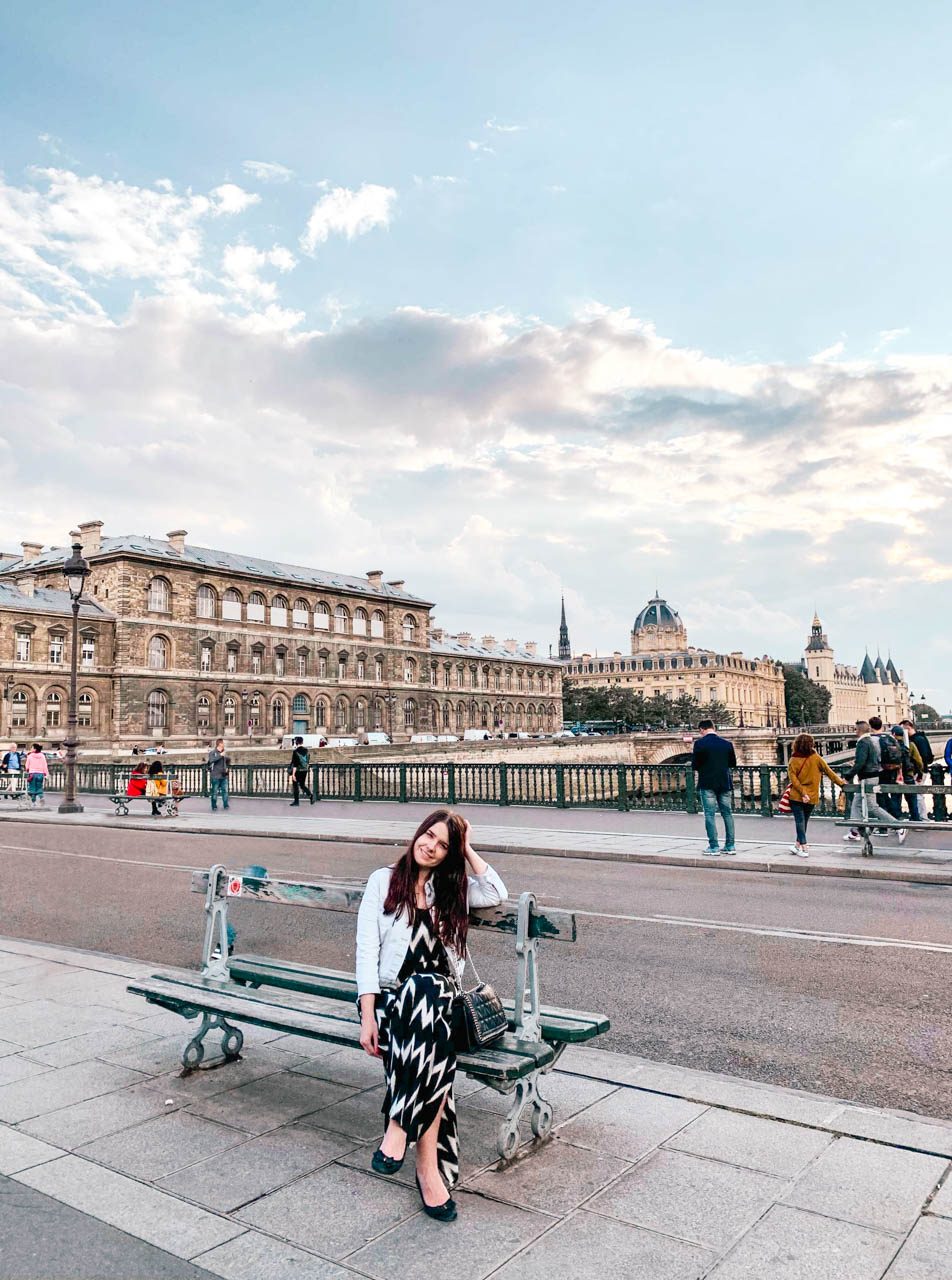 Woman in a denim jacket and a black and white maxi dress sitting on a bench on a bridge in Paris