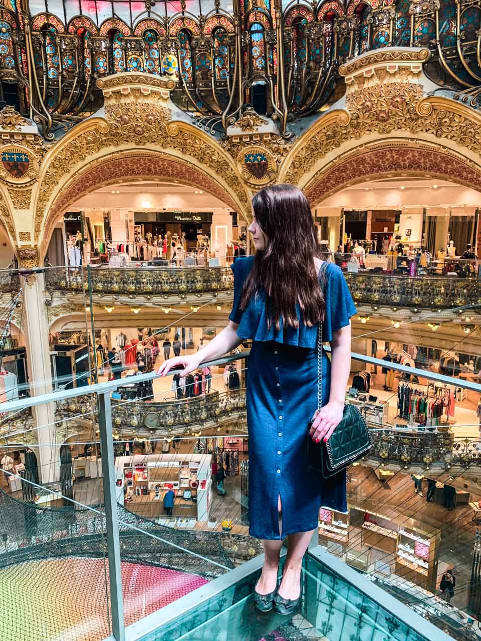 Woman in a blue dress standing on the suspended glass walkway at Galeries Lafayette Paris Haussmann
