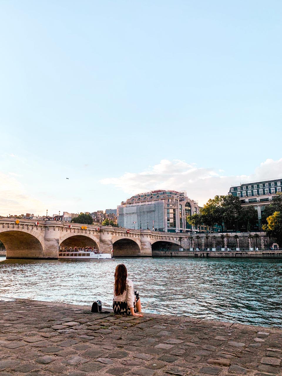 Woman sitting on the bank of the Seine River watching a cruise boat pass under the Pont Neuf