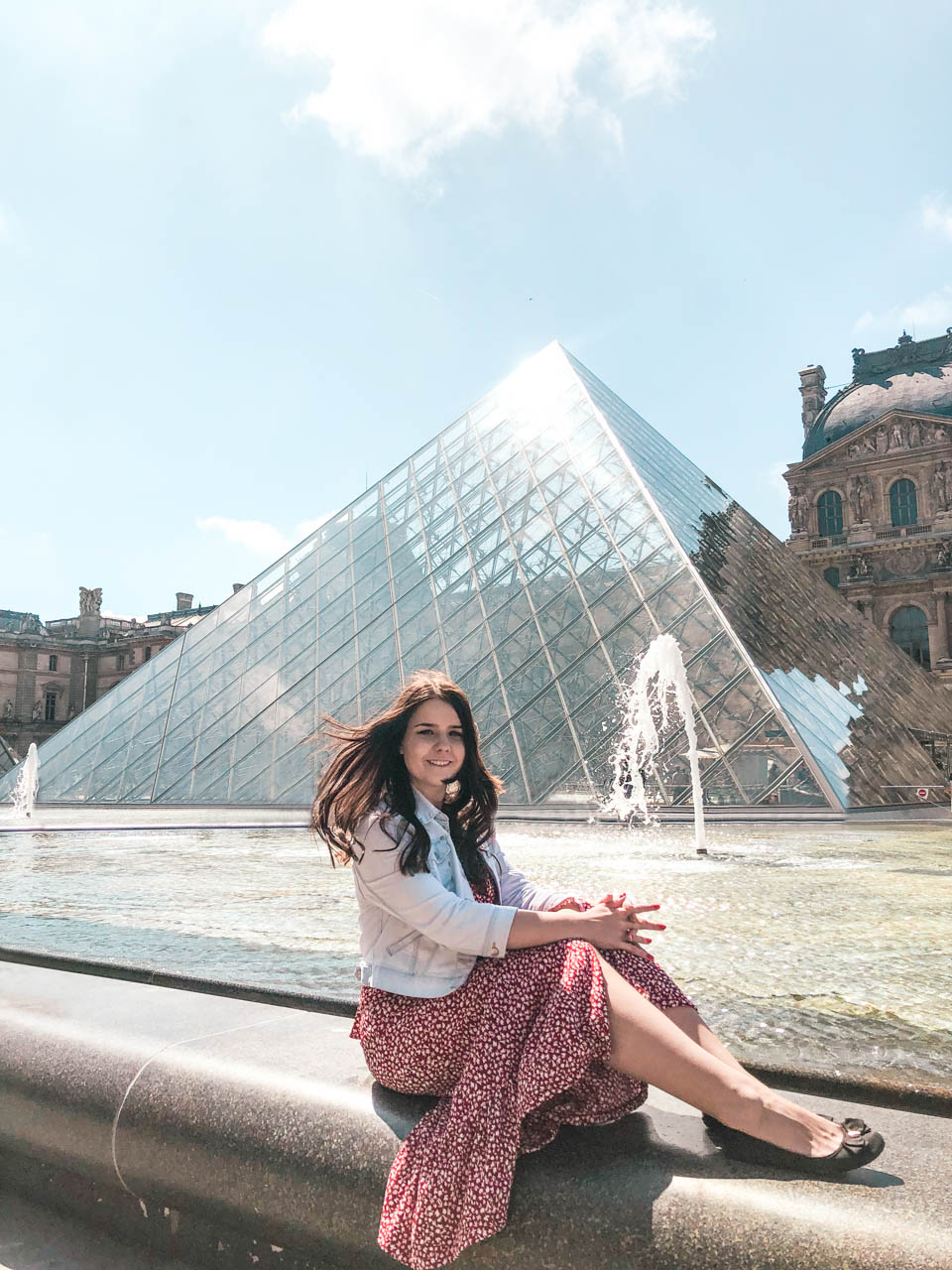 Woman in a red and white floral dress sitting on a fountain in front of the Louvre Pyramid