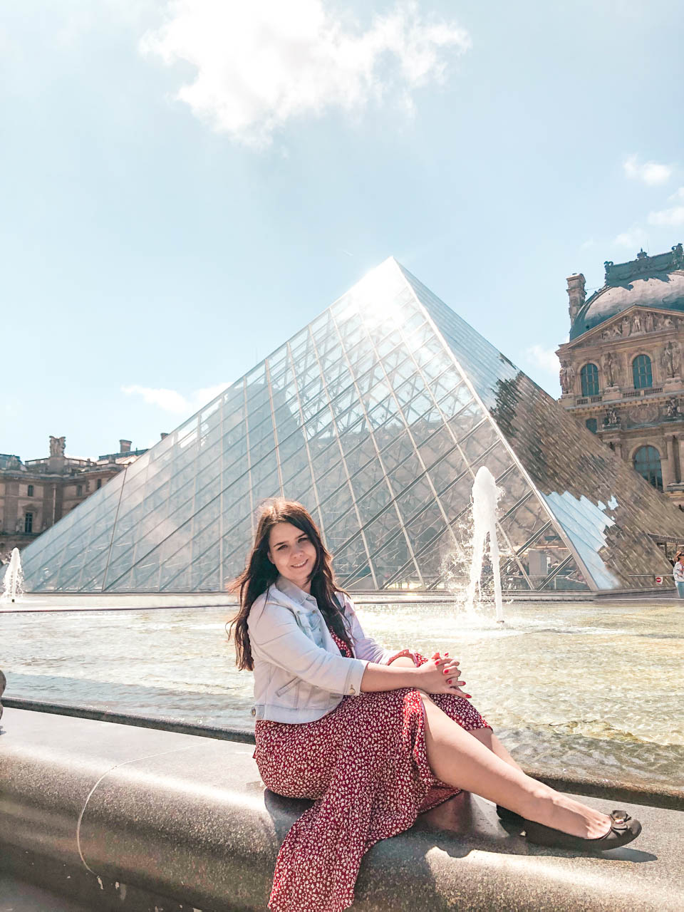Woman in a red and white floral dress sitting on a fountain in front of the Louvre Pyramid
