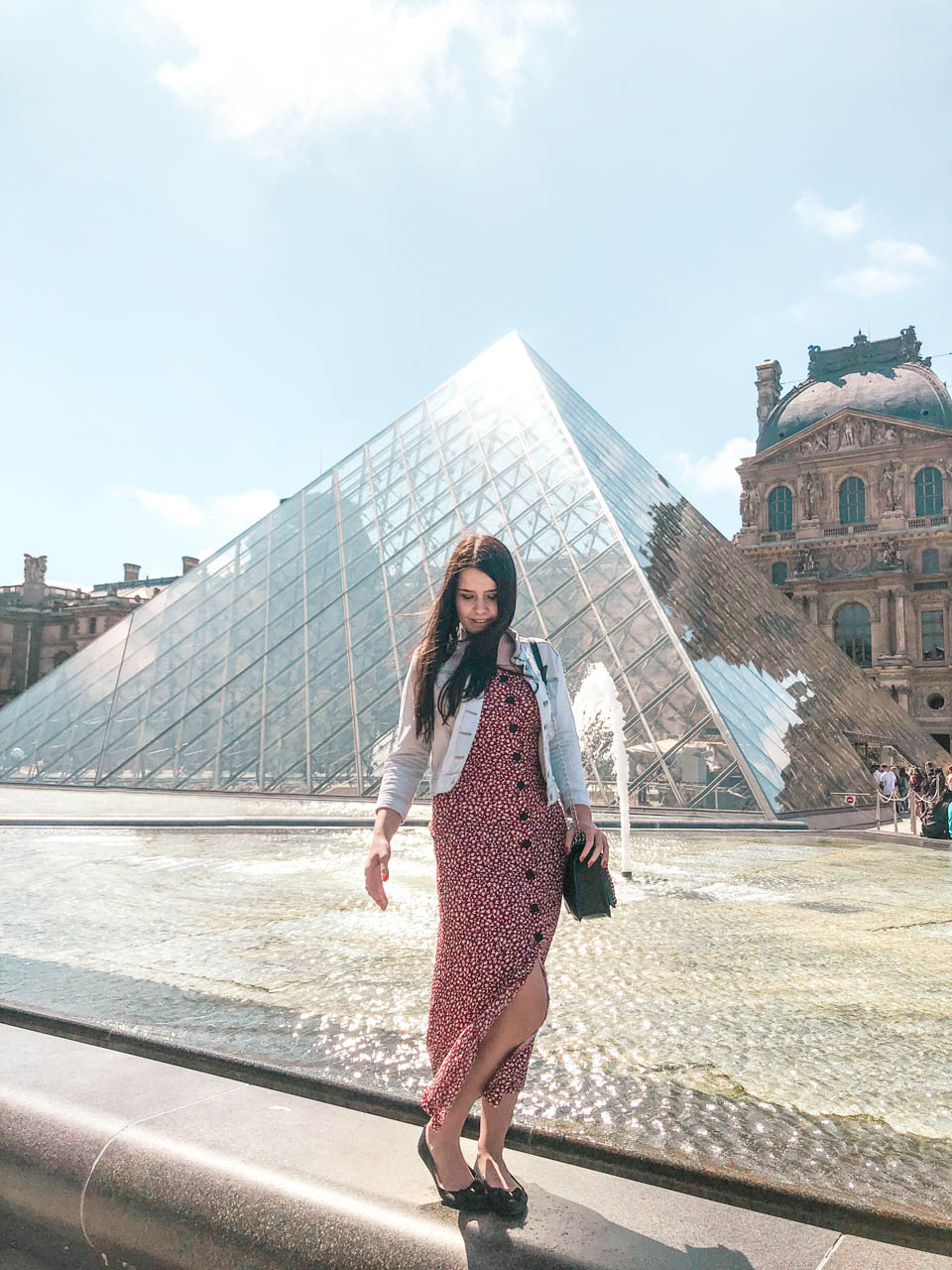 Woman in a red and white floral dress standing on a fountain in front of the Louvre Pyramid