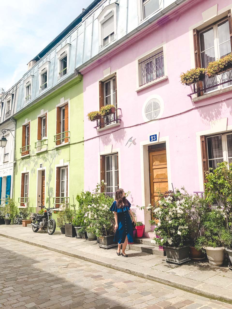 Young dark-haired woman in a blue off-the-shoulder dress looking up at a pink house on Rue Crémieux