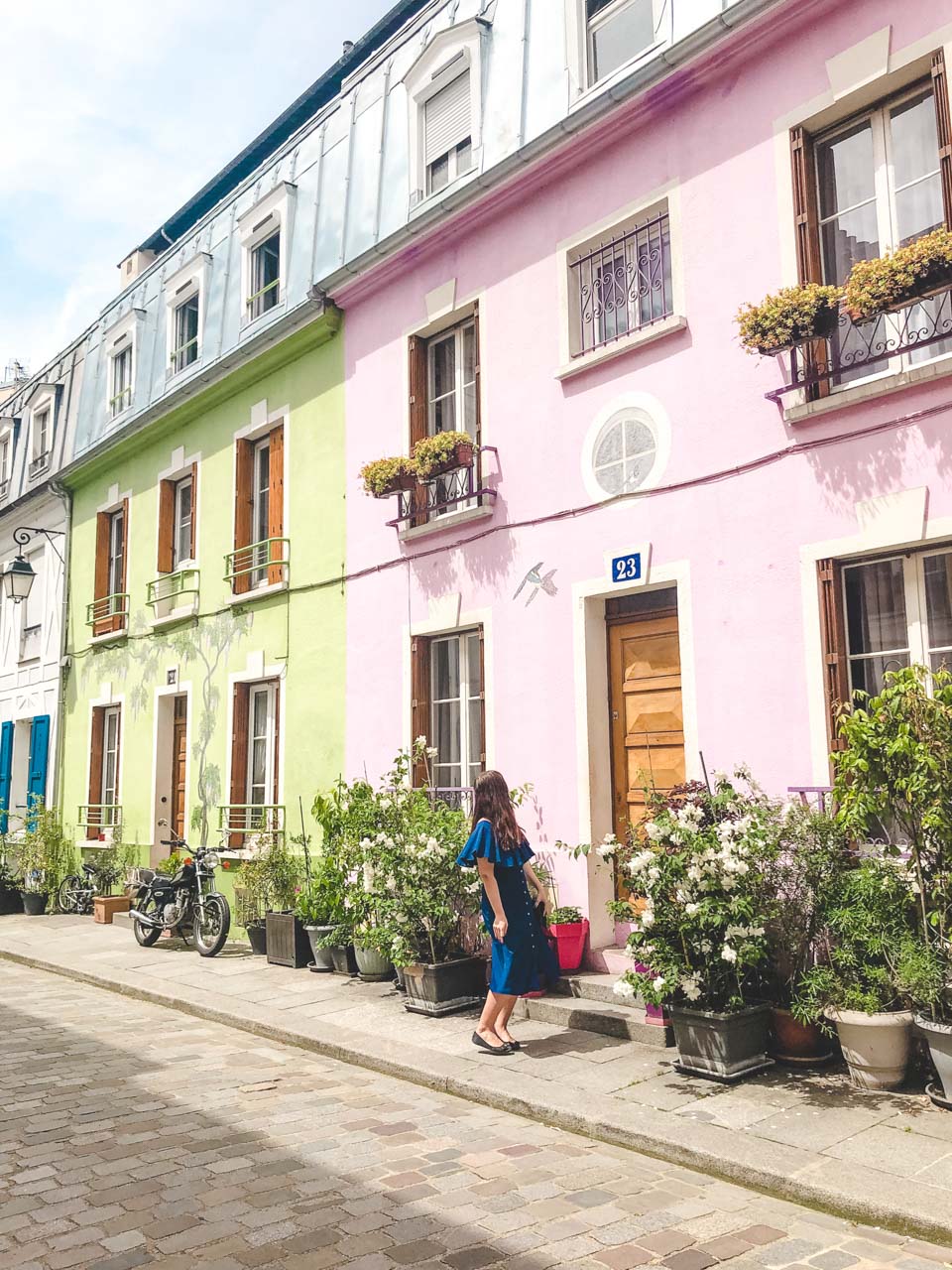 Young dark-haired woman in a blue off-the-shoulder dress looking up at a pink house on Rue Crémieux