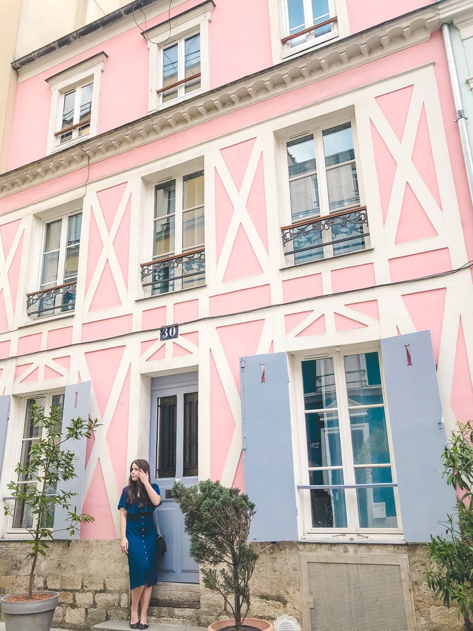 Woman in a blue off-the-shoulder dress standing on the step of a pastel pink house on Rue Crémieux