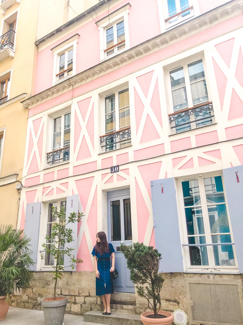 Woman in a blue off-the-shoulder dress standing on the step of a pastel pink house on Rue Crémieux
