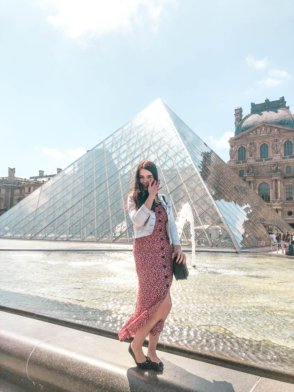 Woman in a red and white floral dress standing on a fountain in front of the Louvre Pyramid