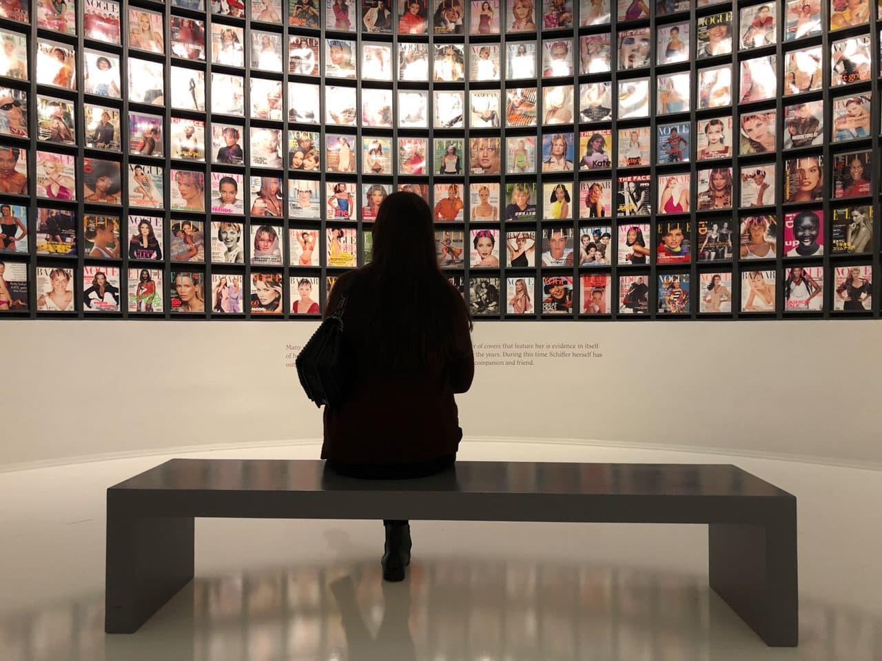 A woman sitting on a bench looking at a wall of framed old magazine covers