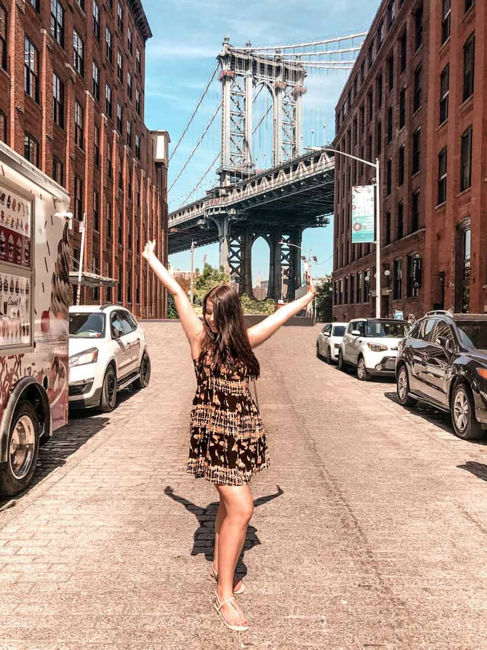 A happy girl in a black patterned dress throwing her hands up in Dumbo, Brooklyn