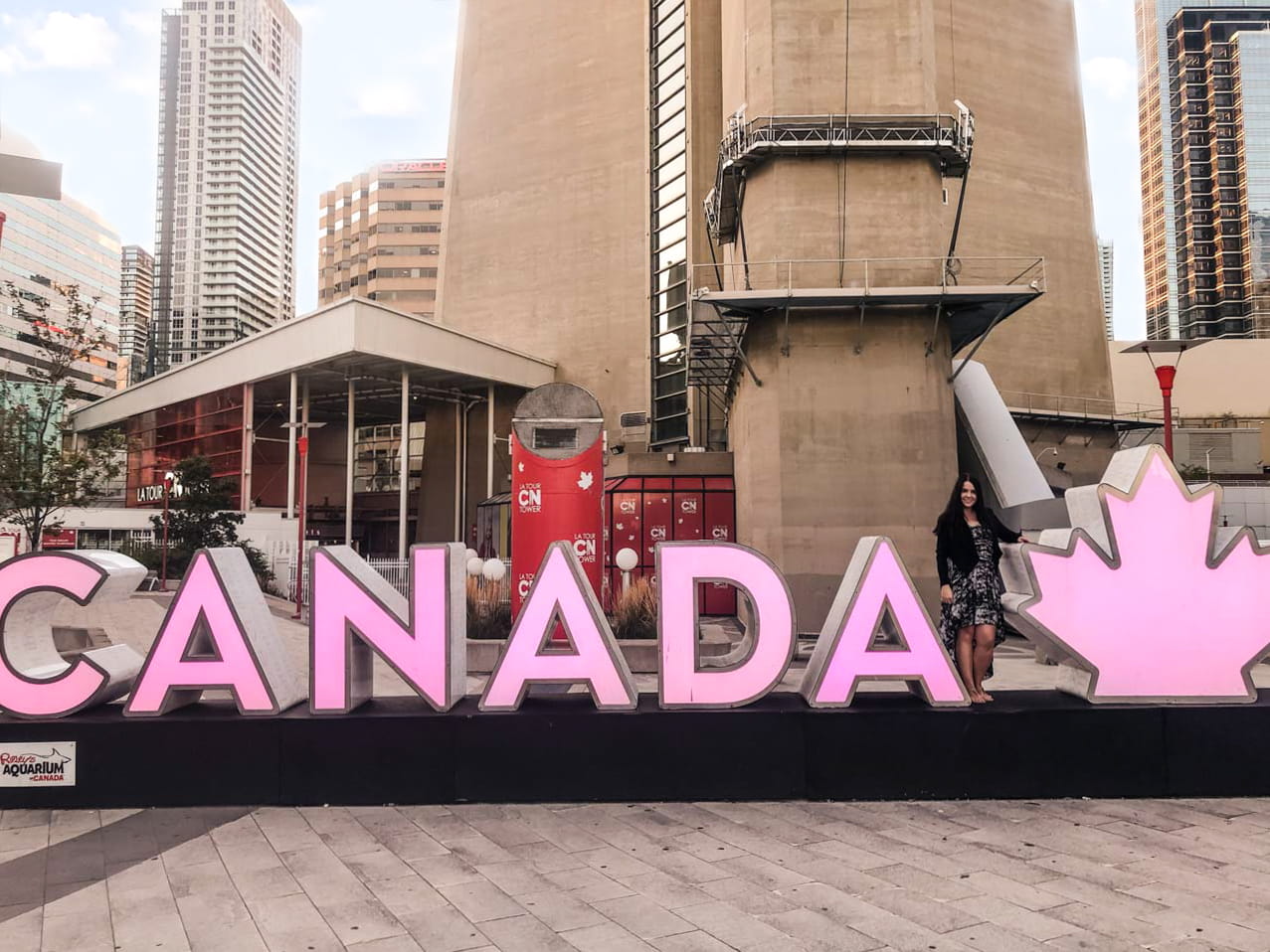 A girl in a maxi dress standing next to a huge Canada sign outside the CN Tower in Toronto