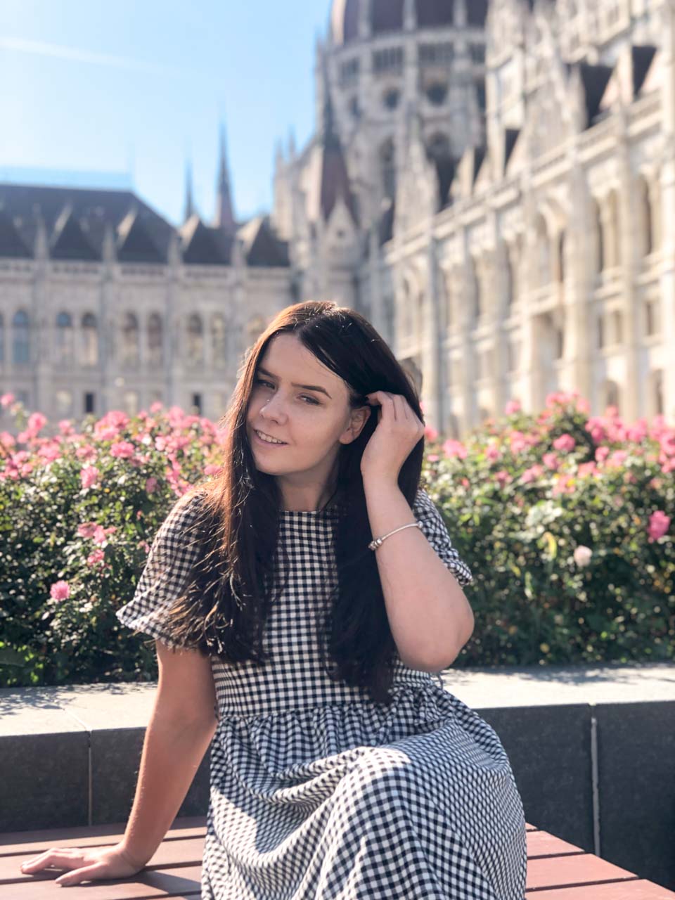 A girl sitting in front of the Hungarian Parliament building, tucking her hair behind her ear