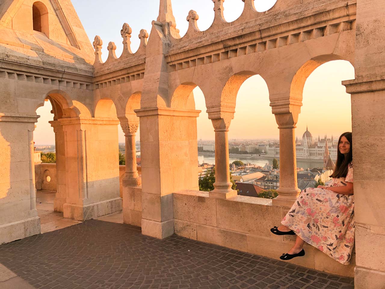 A girl in a maxi dress smiling at the camera as she is sitting on the ledge of the Fisherman's Bastion