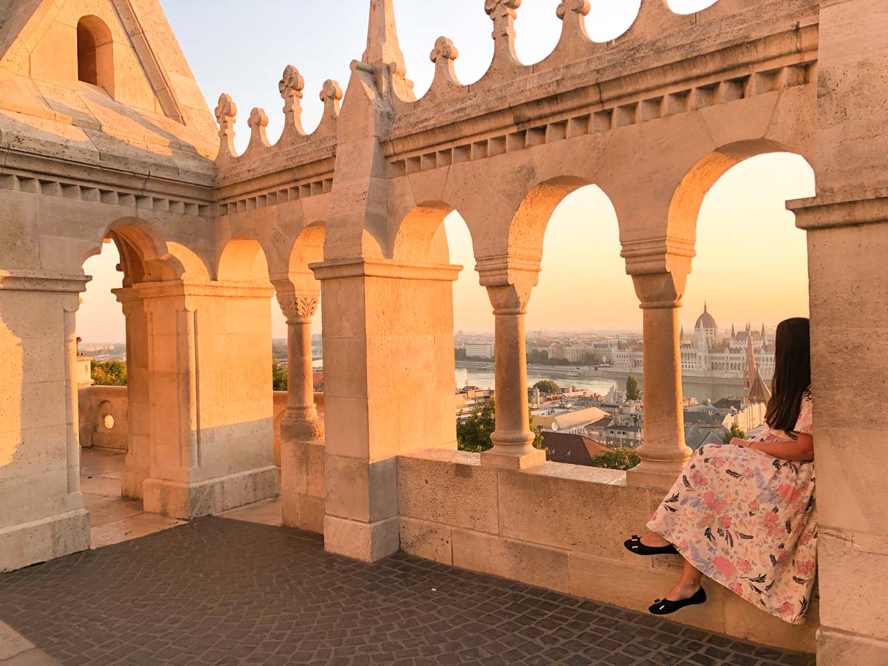 A girl in a maxi dress sitting on the ledge of Fisherman's Bastion looking at the Hungarian Parliament building