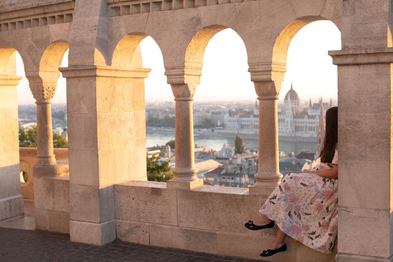 A girl in a maxi dress sitting on the ledge of Fisherman's Bastion looking at the Hungarian Parliament building