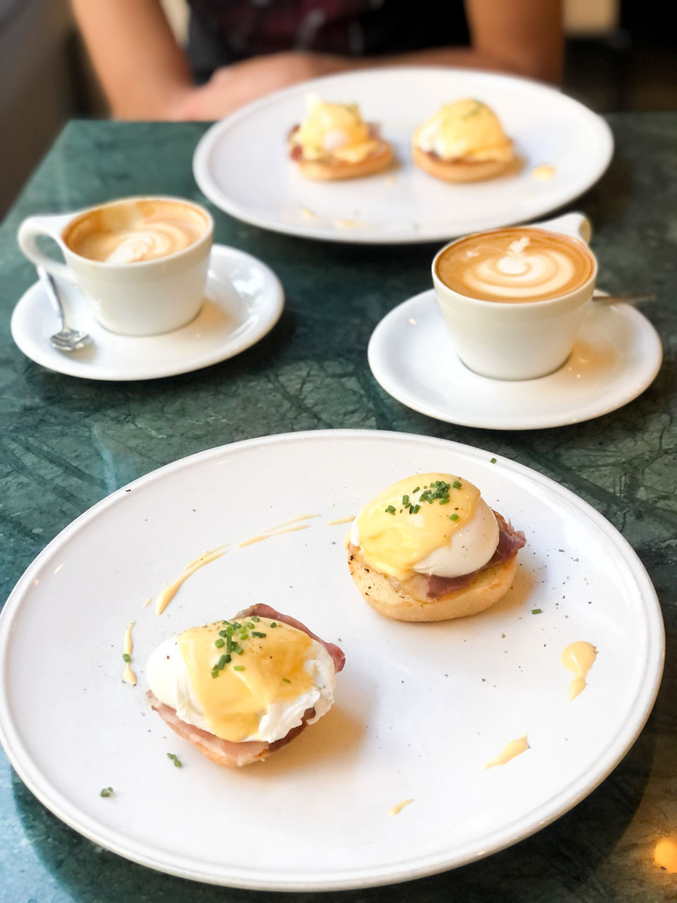Two cups of cappuccino and two plates with eggs benedict on a green stone table
