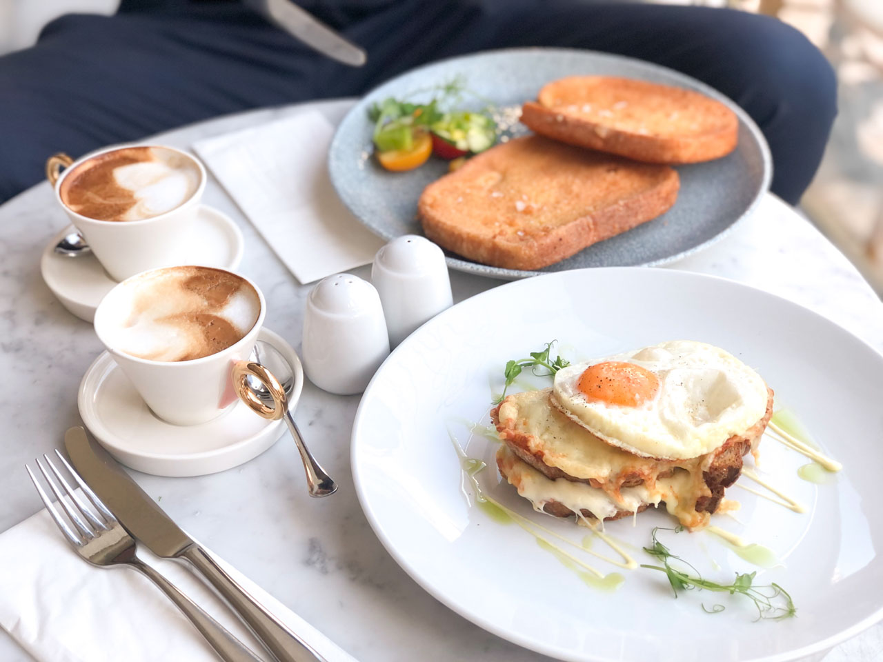 Hungarian French toast on a table next to two cappuccinos and a croque madame sandwich