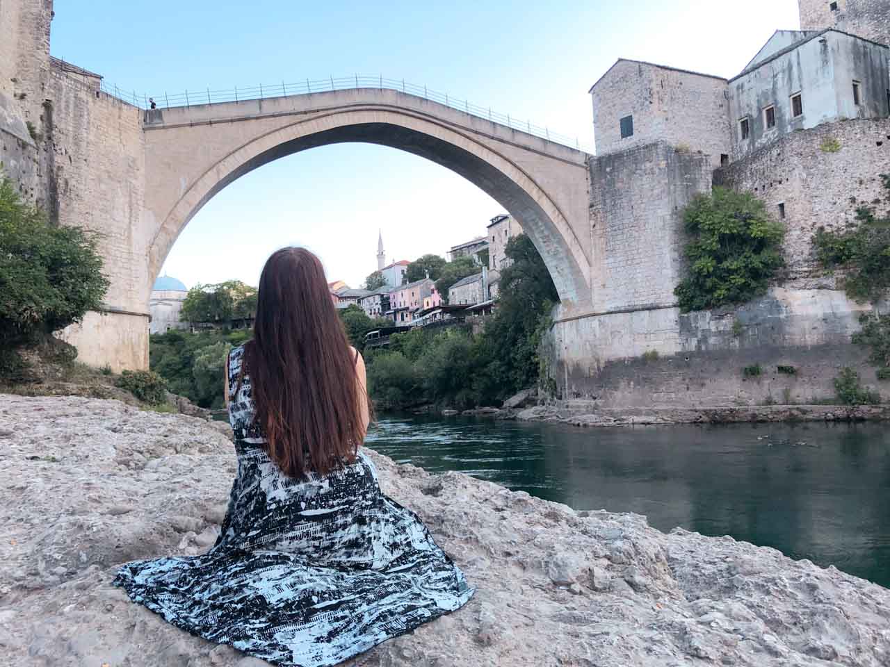 A girl sitting on the beach with her back turned to the camera with Mostar's Old Bridge (Stari Most) in front of her