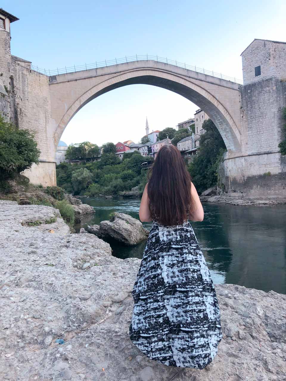 A girl standing on the beach with her back turned to the camera with Mostar's Old Bridge (Stari Most) in front of her