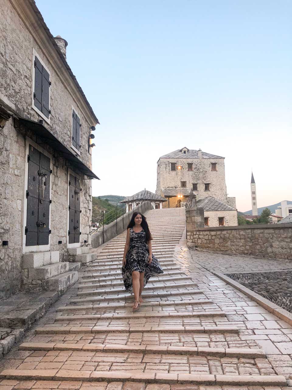 A girl in a black and white maxi dress walking down Mostar's Old Bridge (Stari Most) towards the camera