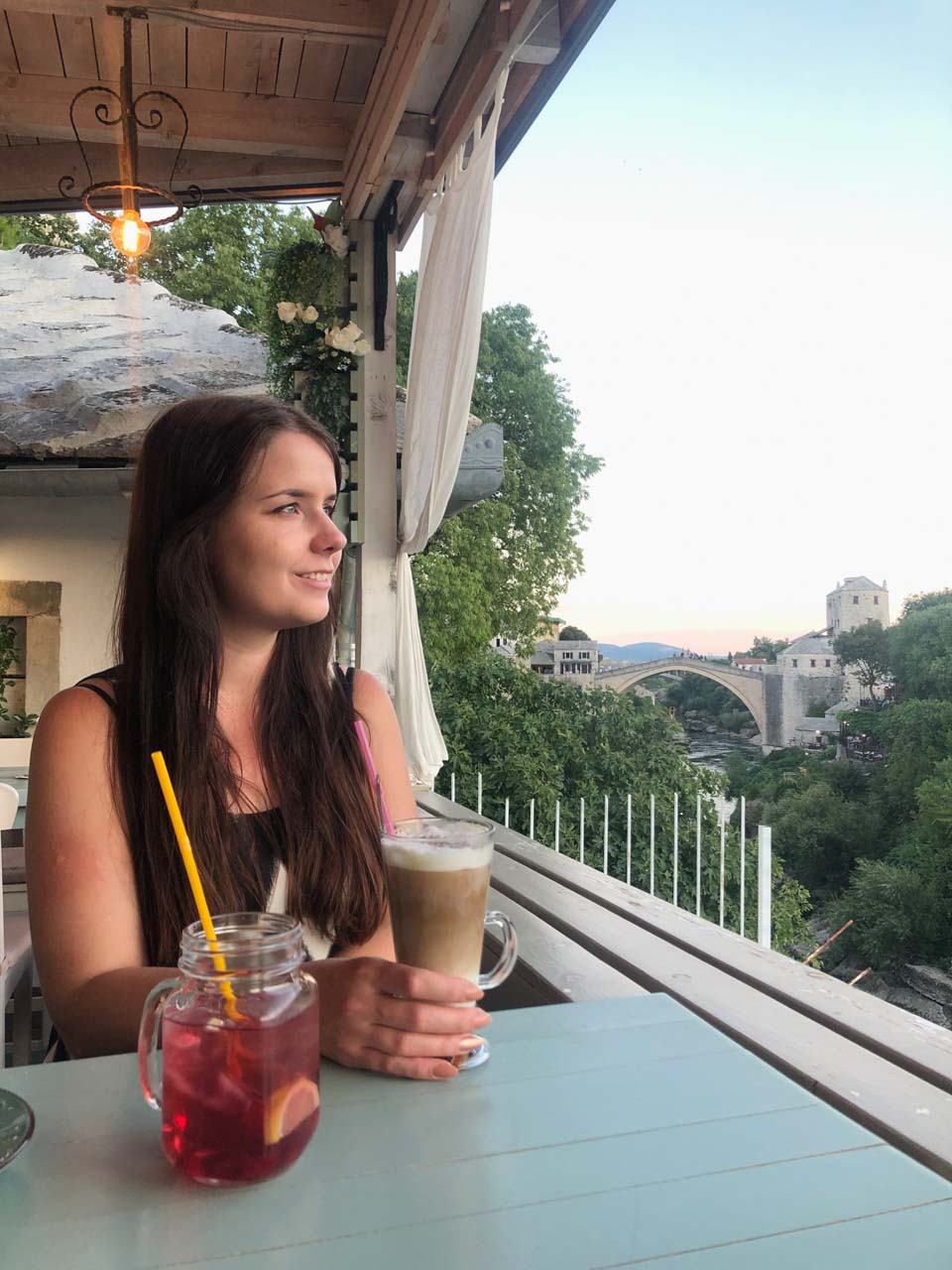 A girl holding a cup of coffee looking sideways with Mostar's Old Bridge behind her