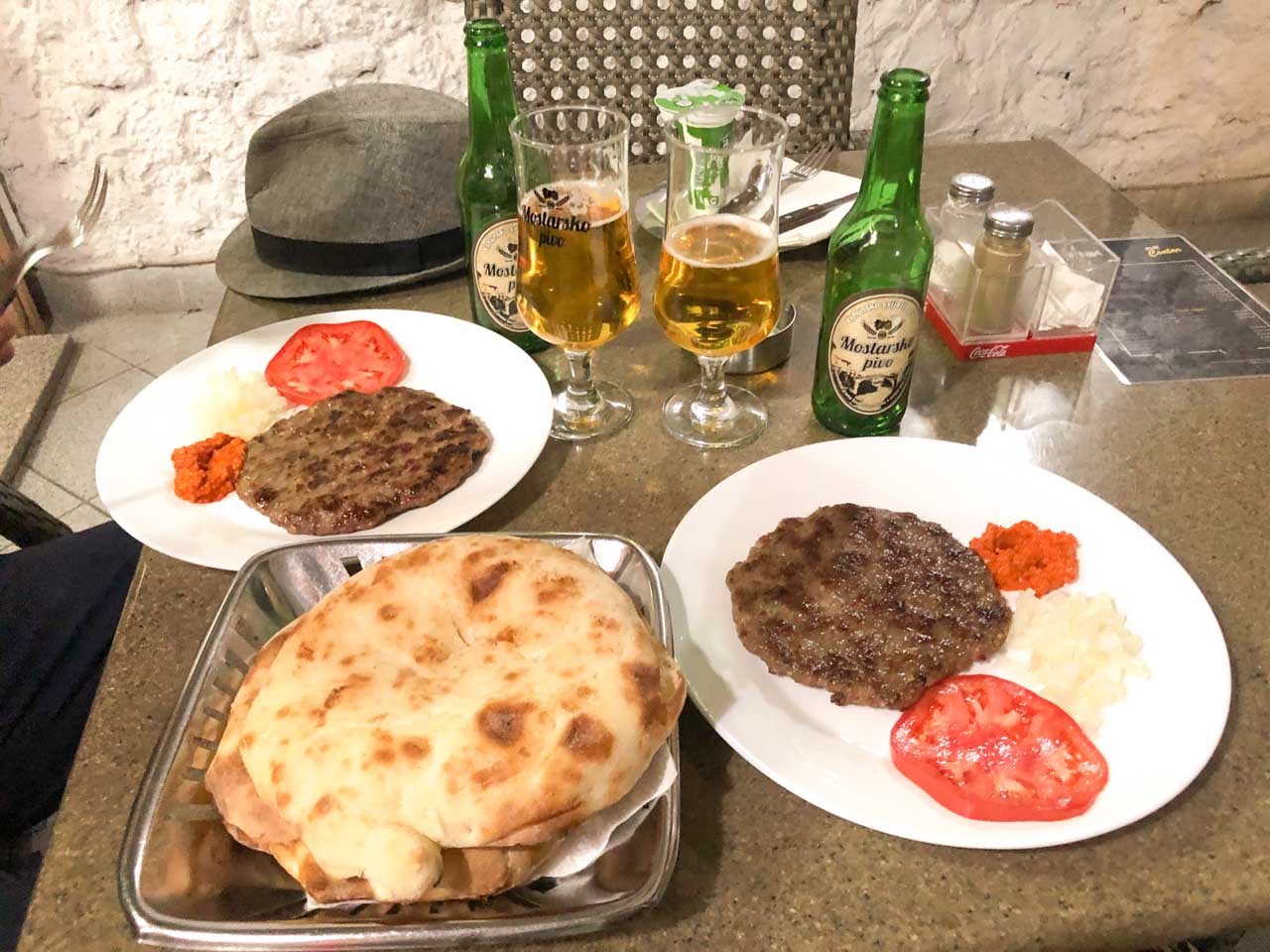 Two plates containing pljeskavica with sides next to pita bread and two glasses of beer on a table