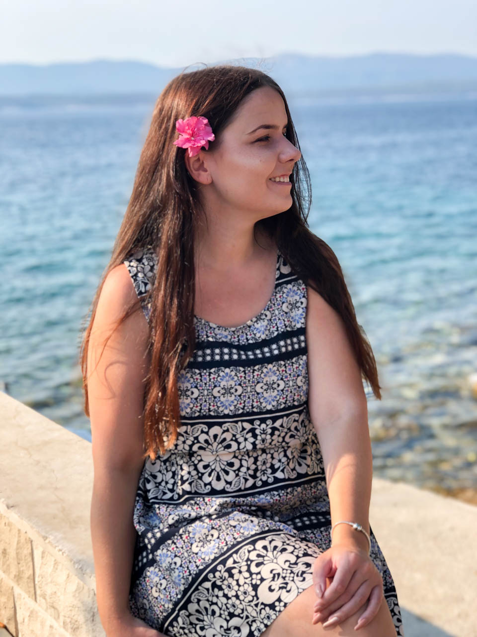 A smiling girl in a summer dress with a pink flower in her hair sitting on top of a stone wall with the Adriatic Sea behind her