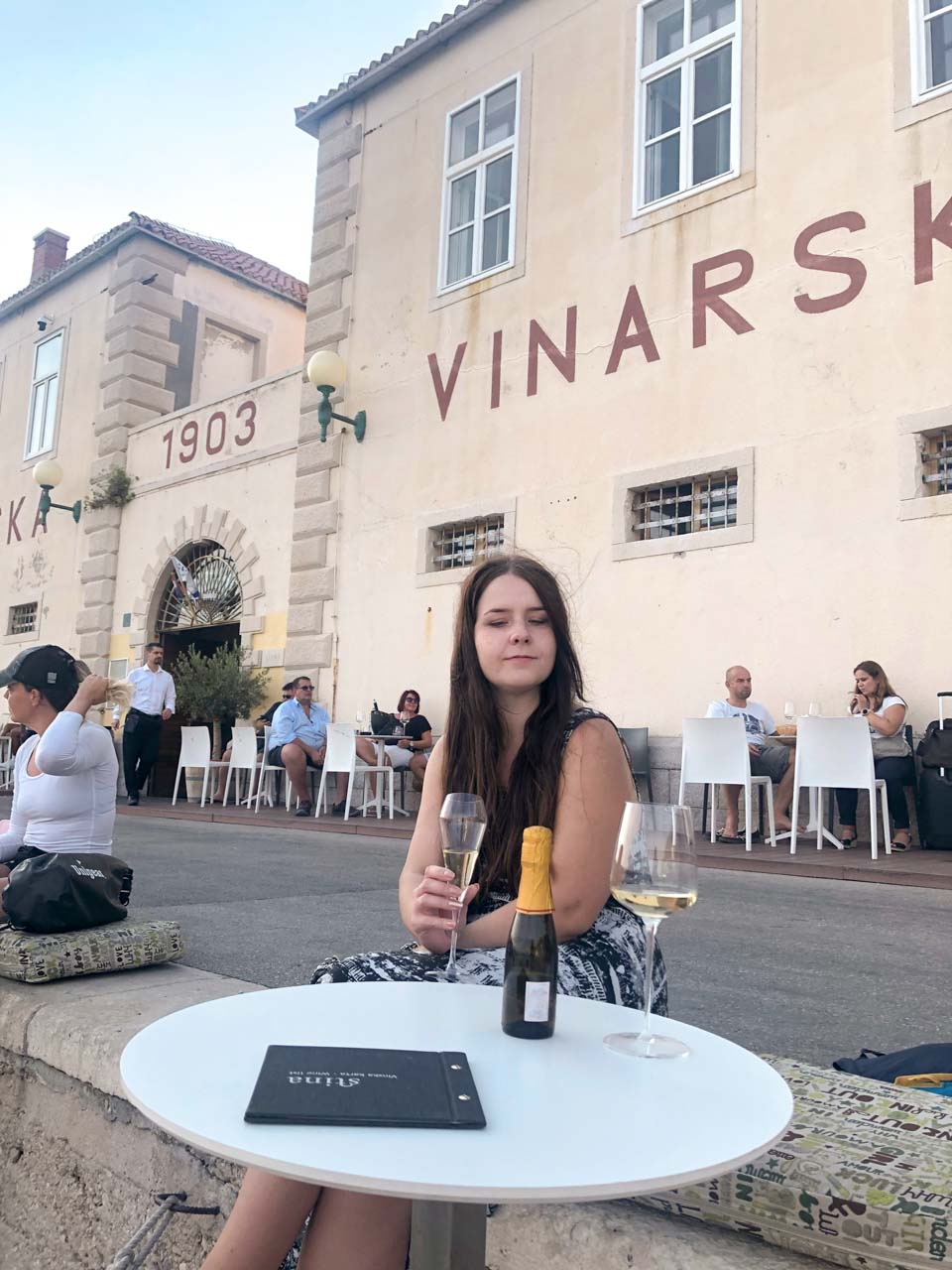 A girl sitting on top of a stone wall with a glass of sparkling wine in her hand with the Stina Winery behind her